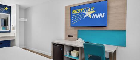 A hotel room with a bed, a desk with a chair, a small refrigerator, a wall-mounted TV displaying 'BESTSTAR KINN', and a mirror with toiletries.