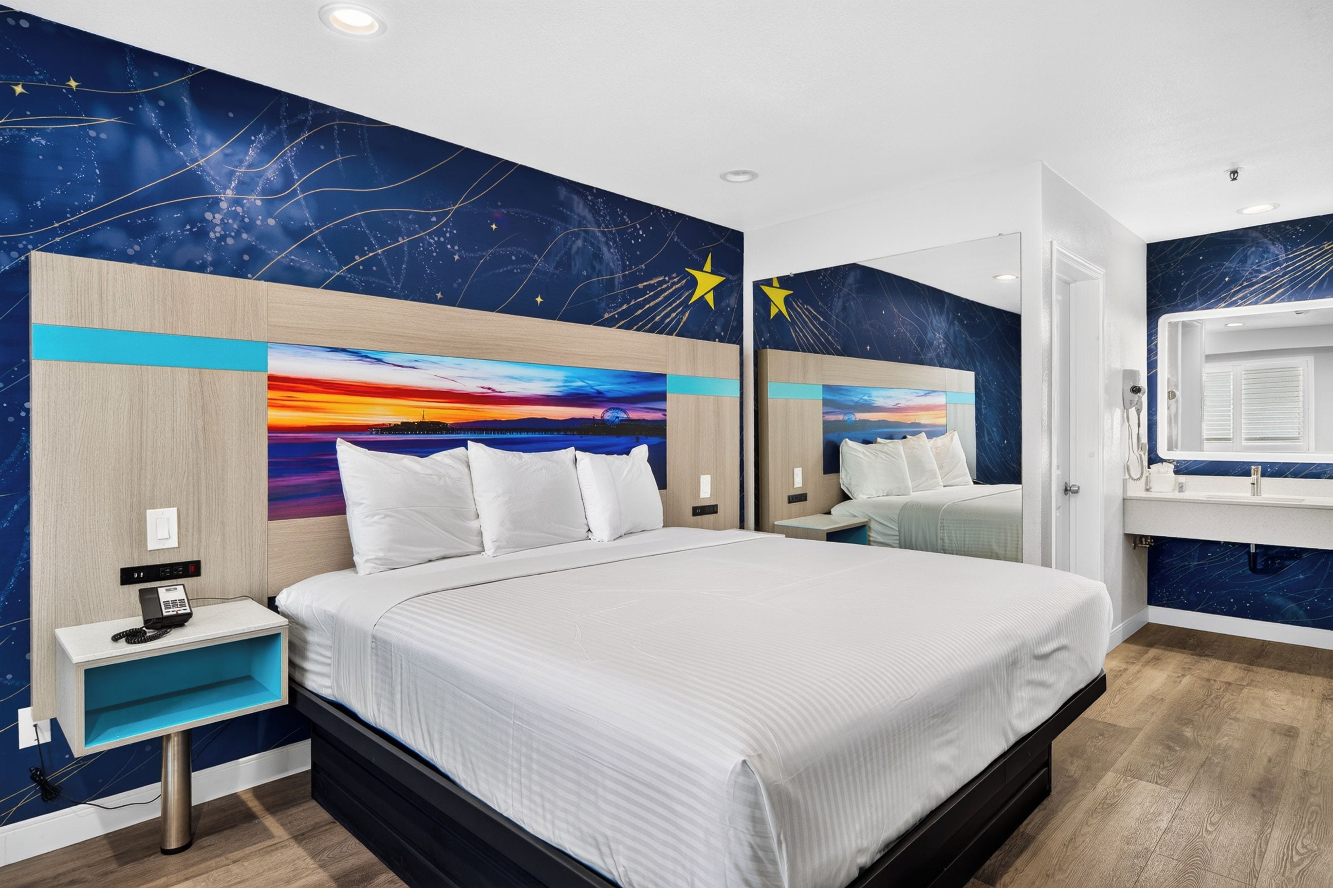 Een hotelkamer met een groot bed, een nachtkastje met een telefoon, een spiegel en een nachtkastje. De muren zijn versierd met een motief van een sterrenhemel.