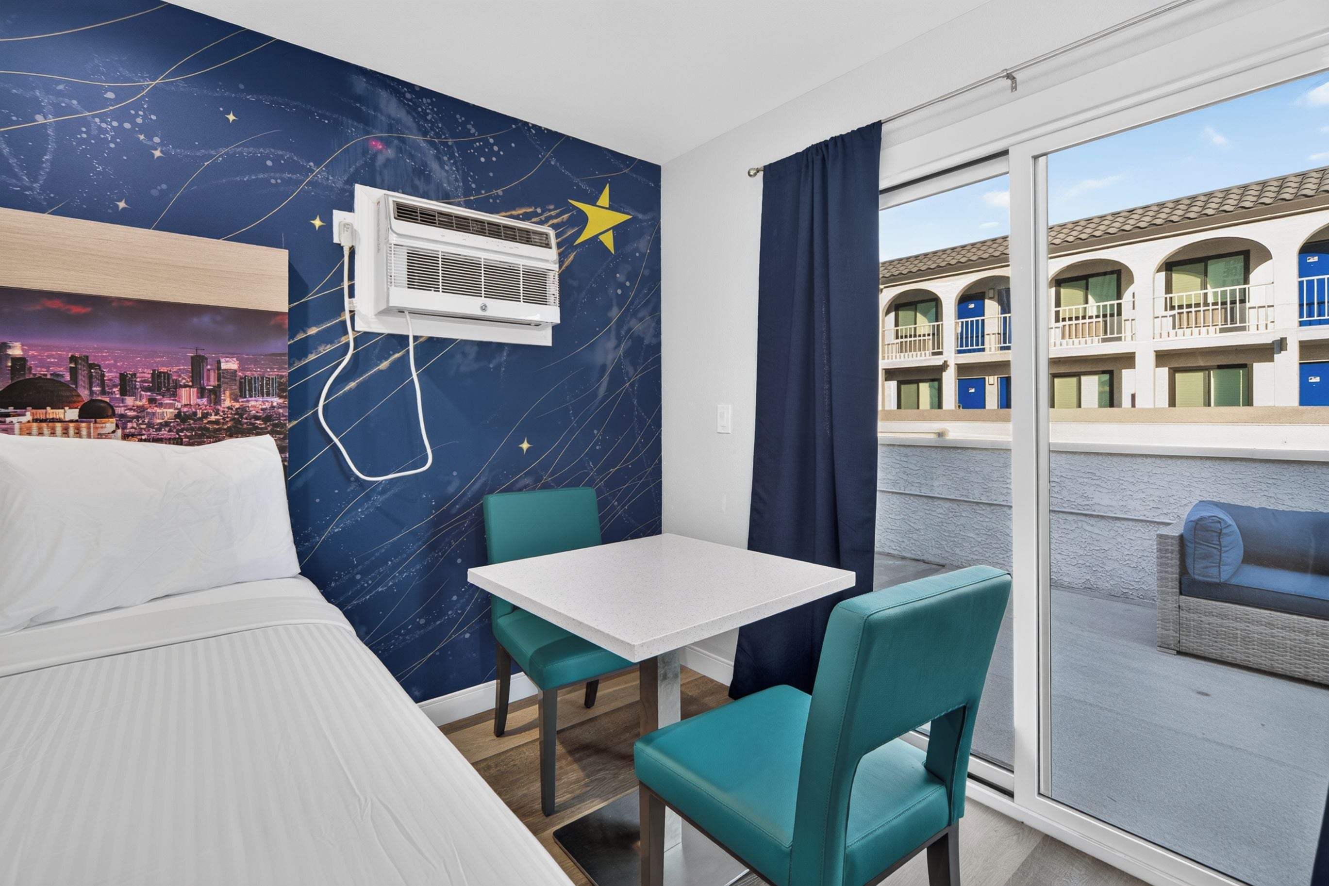 Habitación de hotel con cama, una mesa, dos sillas, un ventanal con vista a un edificio y una pared con un mural de un paisaje urbano.