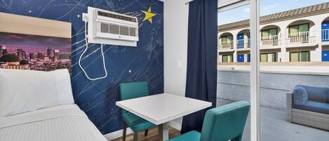 Habitación de hotel con cama, una mesa, dos sillas, un ventanal con vista a un edificio y una pared con un mural de un paisaje urbano.