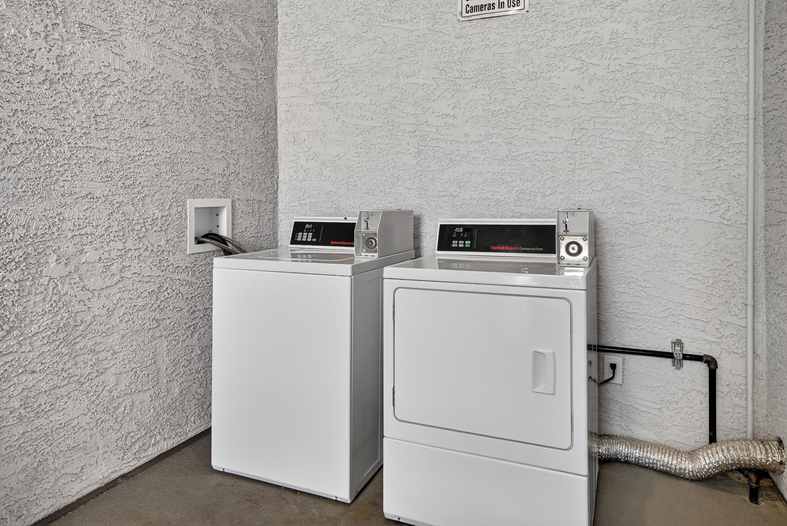Two front-loading washing machines in a utility room with a textured wall and a sign indicating 'Cameras In Use'.