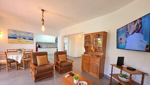 A spacious living room with a dining area, wooden furniture, a kitchen in the background, and a TV mounted on the wall.