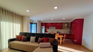 A modern living room with a sofa and ottoman, a dining area with a table and chairs, and a kitchen with red cabinets and a refrigerator.