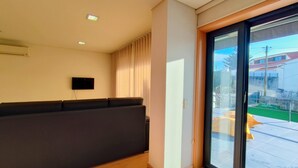 A modern living room with a dark sofa, a flat-screen TV mounted on the wall, and a sliding glass door leading to a balcony.