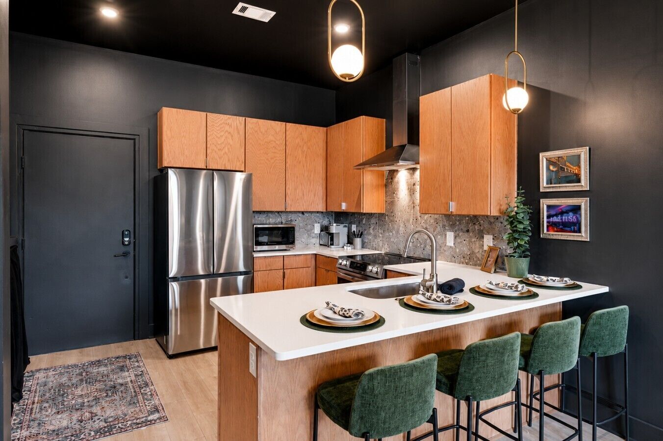 A modern kitchen with a stainless steel refrigerator, wooden cabinets, a central island with bar stools, and pendant lighting.