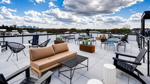 En tagterrasse med udendørs møbler, herunder sofa, stole og borde, samt udsigt over byens skyline i baggrunden.
