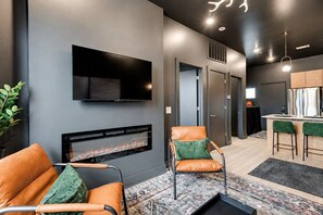 A modern living room with a fireplace, a flat-screen TV mounted on the wall, and a seating area with two chairs. - East Nash Top-Floor 1BR | Rooftop Deck | Sleeps 6 (Nashville)