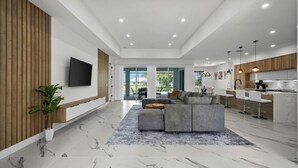 A spacious living area with a large sectional sofa, a flat-screen TV mounted on the wall, and a kitchen area with modern appliances and pendant lighting. - Luxury 3-bedroom Villa with Heated pool and spa (Rotonda West)