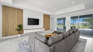 A modern living room with a grey sectional sofa, a flat-screen TV mounted on the wall, and a large sliding glass door leading to a patio. - Luxury 3-bedroom Villa with Heated pool and spa (Rotonda West)