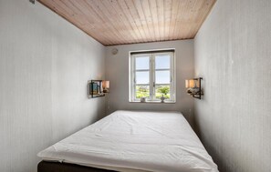 Een smalle kamer met een houten plafond, een raam met uitzicht op groen, twee wandlampen en een bed met een wit laken.