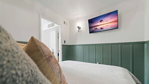 A bedroom with a bed, a green headboard, a painting of a sunset, and a door leading to another room. - Sunset Cottage (Fairfield Bay)