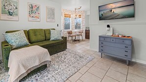 A living room with a green sofa, patterned throw blanket, and a large rug. There is a TV mounted on the wall and a dresser with drawers. - Sunset Cottage (Fairfield Bay)
