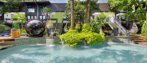 A pool area with a wooden deck, a large spherical mirror, and a staircase leading to a building with a green facade.