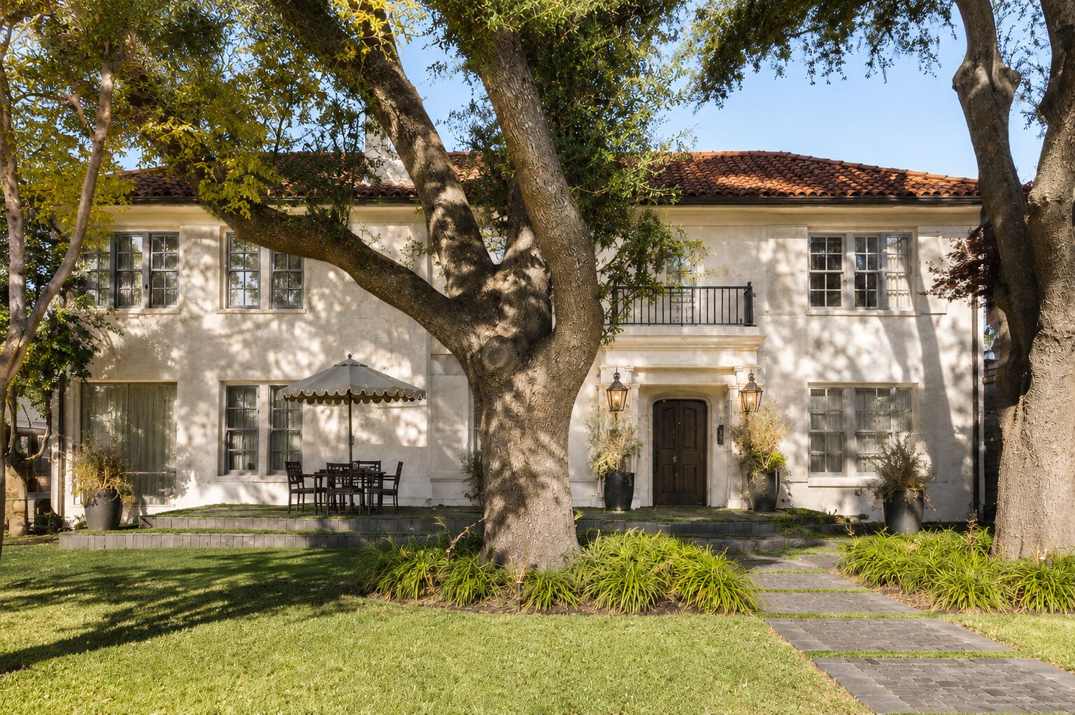 A two-story house with a large tree in the front yard, a patio with a table and chairs, and a well-maintained lawn.