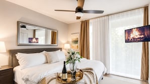 Una habitación de hotel con cama, ventilador de techo, televisión, mesita de noche, cómoda y ventana con cortinas.
