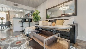 A modern living room with a dark leather sofa, a round coffee table, and a large mirror above the sofa. There is a dining area with a bar and two black bar stools. A potted plant is on the sofa, and a chandelier hangs from the ceiling. - 2-level Spacious loft   with pool, private patio, and historic charm. (Dallas)