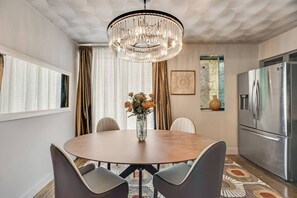 Un comedor con una mesa redonda, dos sillas, un florero con flores, una araña, un refrigerador y una ventana con cortinas.