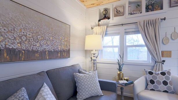 Een woonkamer met een grijze bank, een witte fauteuil en een kamerbreed tapijt. Er hangt een schilderij aan de muur, en er staat een lamp. Een raam met gordijnen is ook te zien.