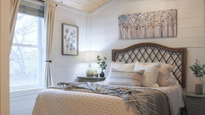 Een slaapkamer met een groot bed, een nachtkastje met een lamp, een aan de muur hangend schilderij en een raam met gordijnen.