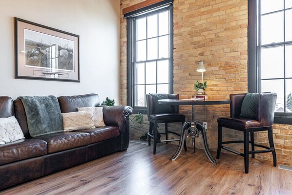 A living room with a leather sofa, a dining table with chairs, a lamp, and a large window with a view of the outside.
