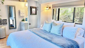 A bedroom with a large bed, a bedside table, a mirror, a window with blinds, and a view of greenery outside. - Peaceful LA Retreat w/ King Bed, Hot Tub and Mountain Views! Enjoy the Outdoors (Topanga)