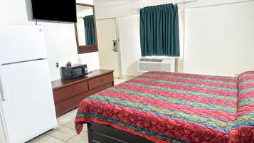 Un dormitorio con cama, televisor, refrigerador, microondas, cómoda y ventana con cortinas.