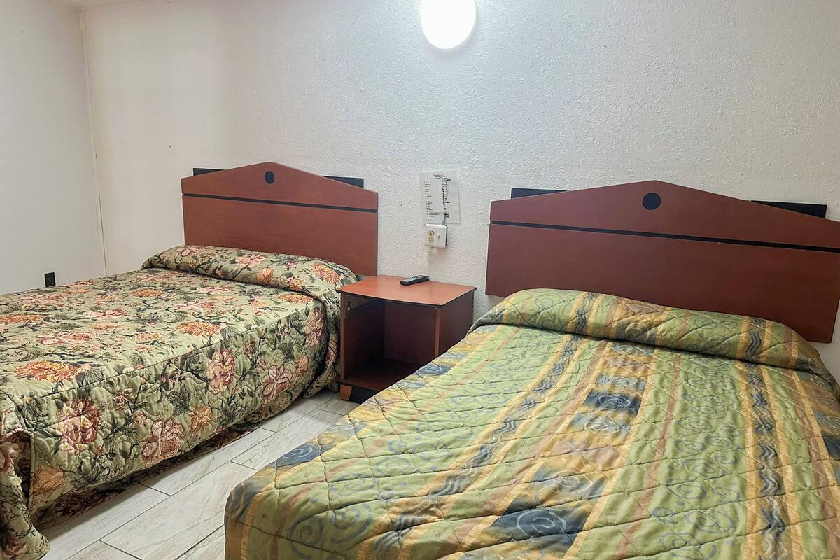 A room with two beds, each with a wooden headboard and floral bedding. There is a nightstand between the beds, and a wall socket is visible.