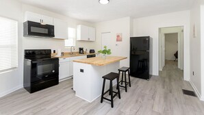 A modern kitchen with white cabinetry, a black stove, a microwave, and a refrigerator. There are two bar stools and a small island with a plant.