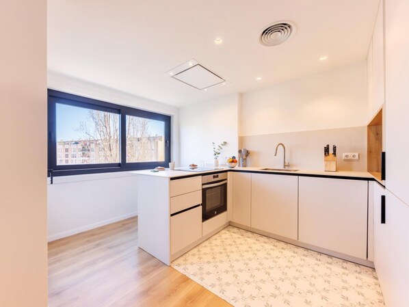 Una cocina moderna con un gran ventanal, muebles blancos, horno integrado y suelo estampado.