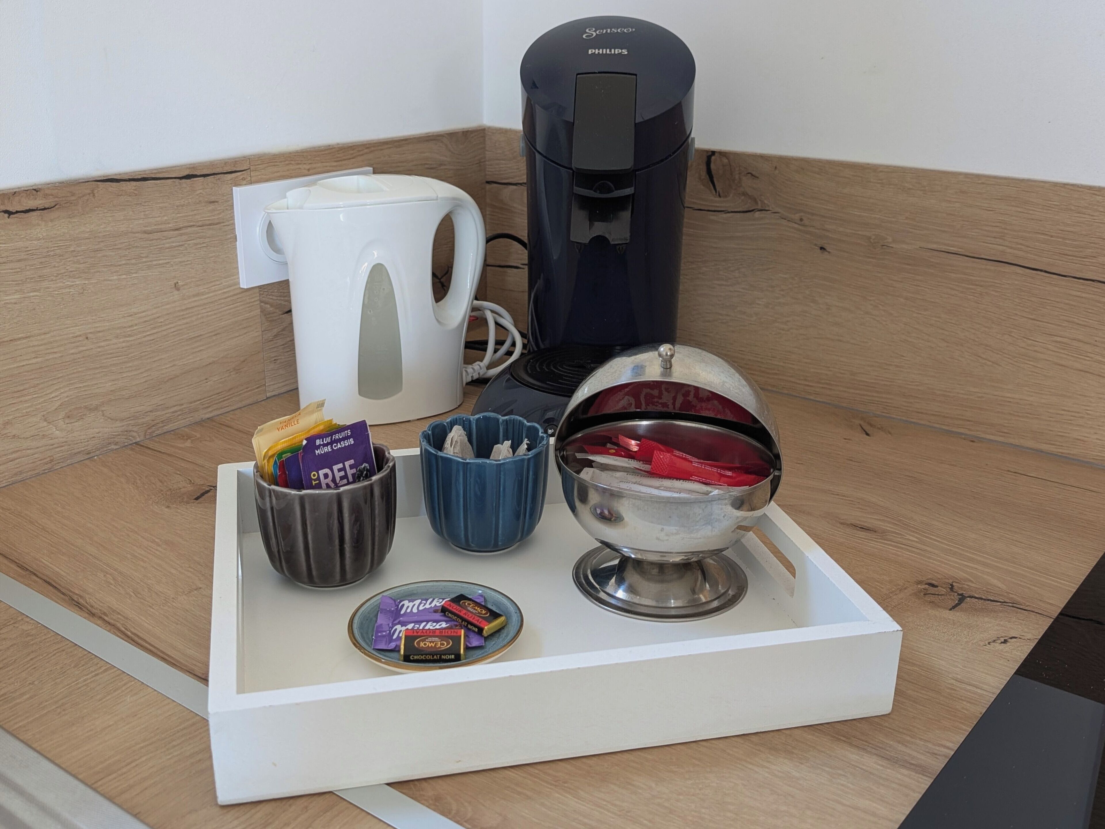 A coffee maker, a white mug, a blue cup, a small bowl with snacks, and a chocolate bar on a tray.