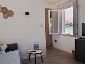 A room with a bed, a small table with a book, a TV, and a window with a view of a body of water.