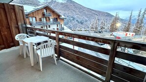 Un balcón de madera con una mesa y sillas, con vistas a un paisaje montañoso con árboles nevados y un edificio al fondo.