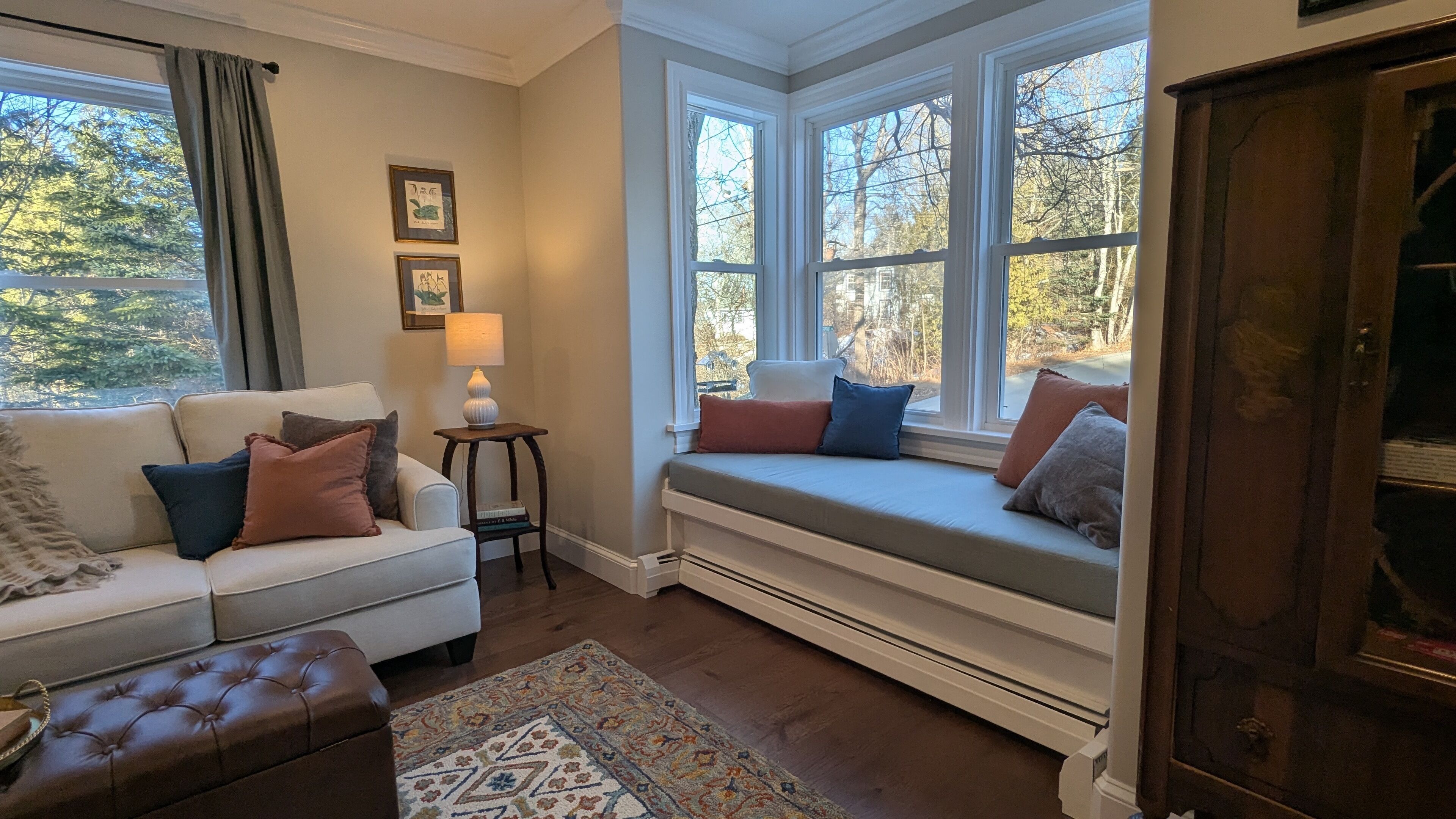 A cozy living room with a white sofa, a bench with cushions, a small table with a lamp, and a view of trees through large windows.