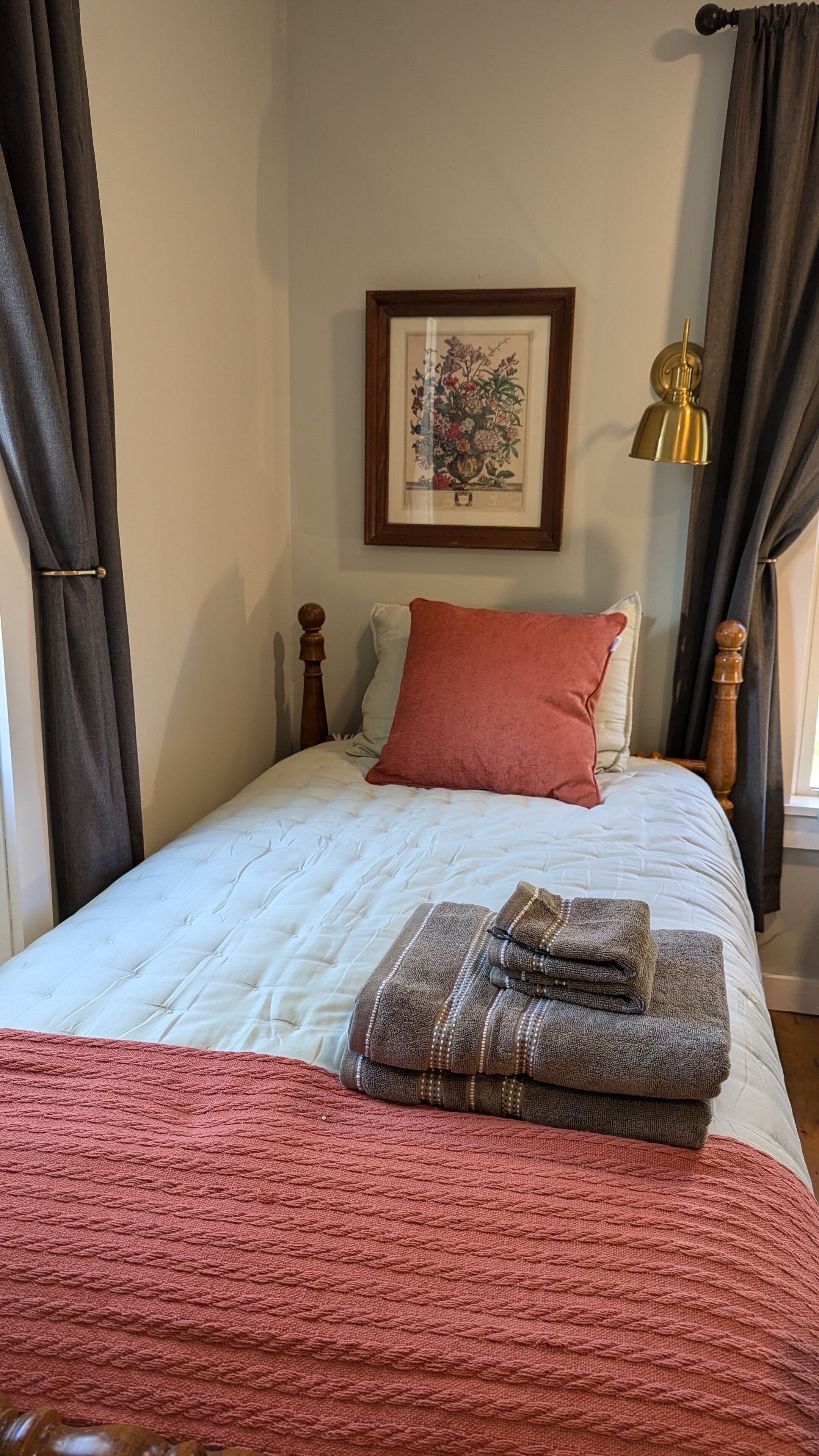 A neatly made bed with a red pillow, a folded towel, and a framed floral artwork on the wall.