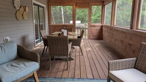 Eine überdachte Terrasse mit Holztisch und -stühlen, umgeben von großen Fenstern mit Blick auf den Wald.