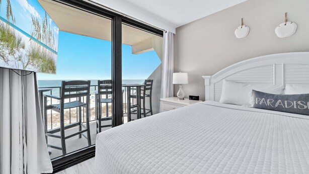 Habitación de hotel con una cama grande, vista a la playa y un balcón con sillas.