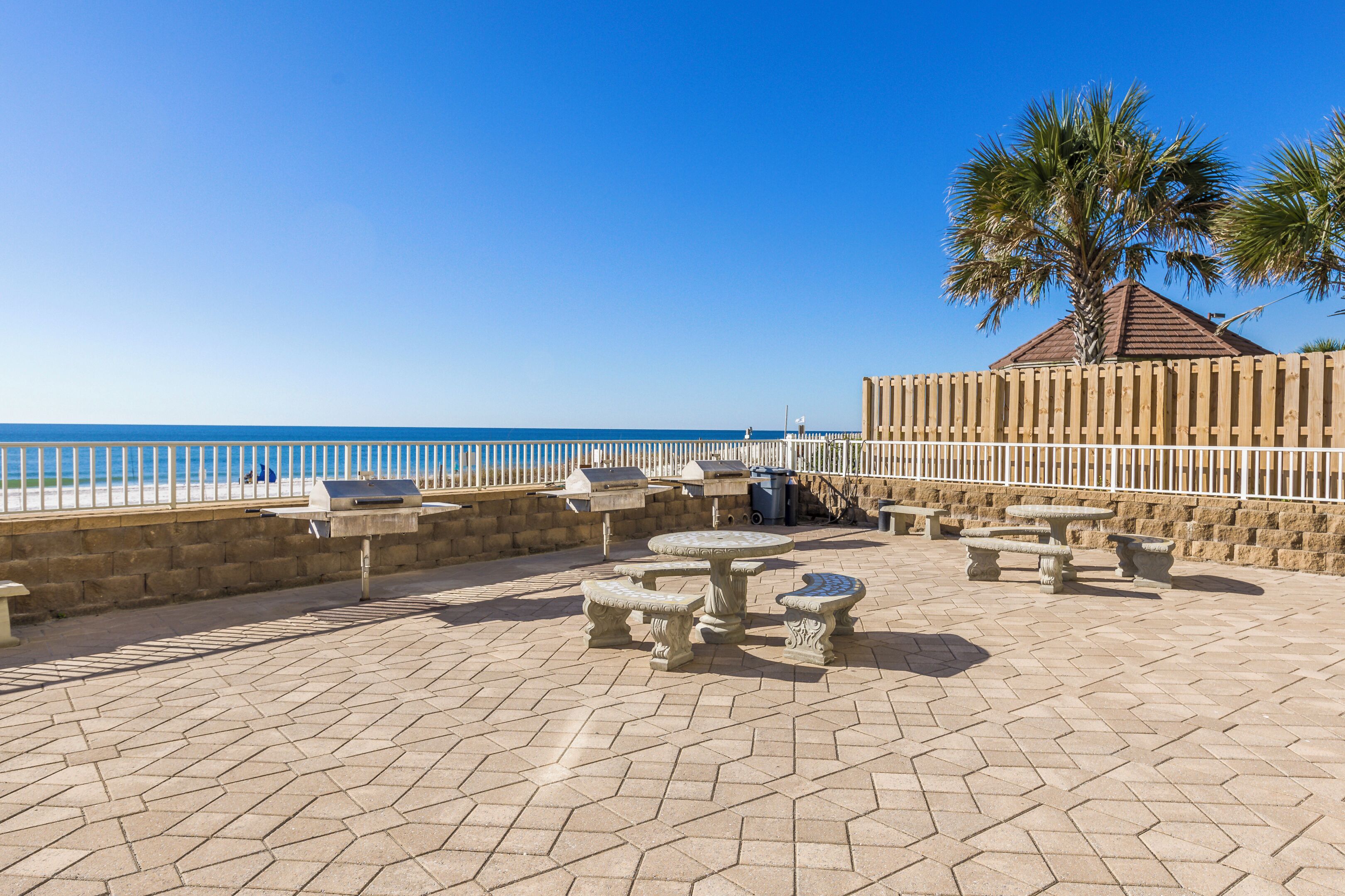 An outdoor patio with stone tables and benches, a palm tree, and a view of the beach.
