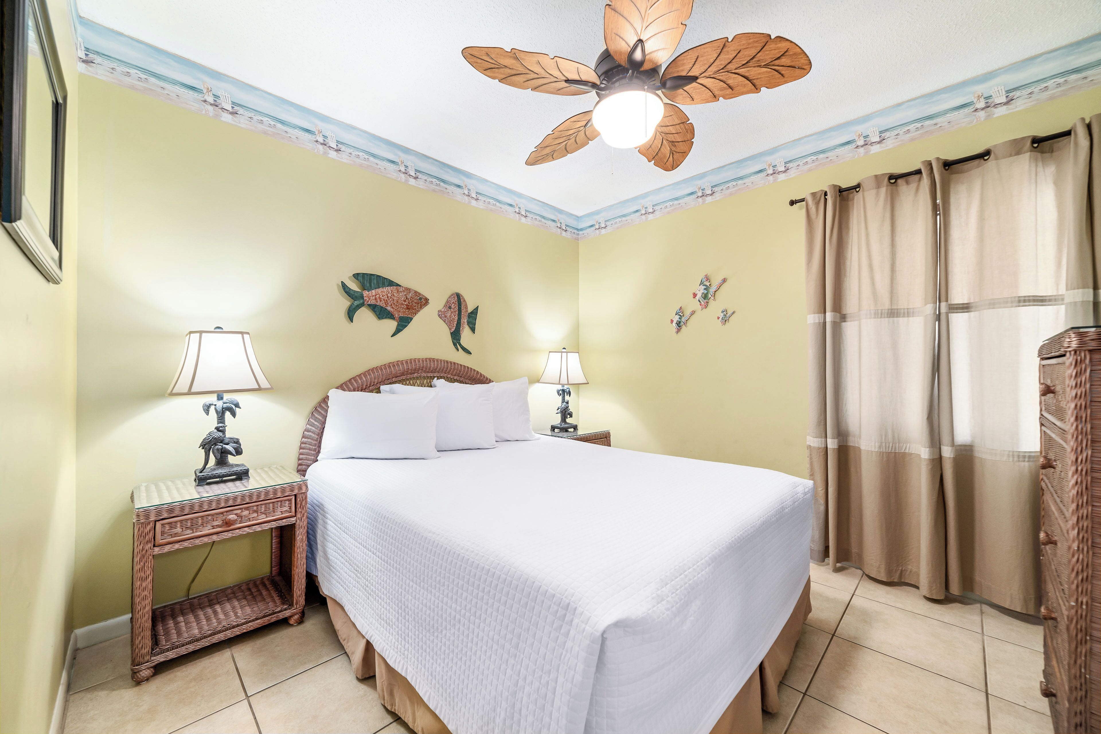 Una cama bien hecha con sábanas blancas, un ventilador de techo, una mesita de noche de mimbre con lámparas y un cuadro decorativo de peces.