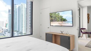 A modern bedroom with a large bed, a TV mounted on the wall, and a view of tall buildings through a large window. - 2409 - Spacious 1BR w Balcony & Stunning Views - Pool, Gym, Near Arena & Bayside (Miami)