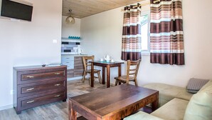 Un salon chaleureux avec une table basse en bois, un canapé et une table à manger avec des chaises. On aperçoit une cuisine à l’arrière-plan, avec un four à micro-ondes et une bouilloire.