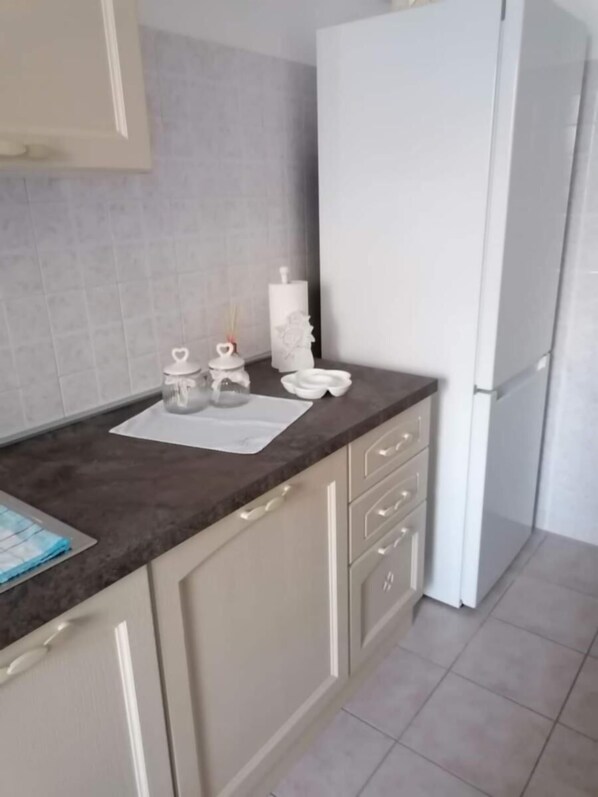 Una cocina con encimera oscura, armarios blancos y un refrigerador blanco. Hay dos recipientes blancos en la encimera, uno con tapa con forma de corazón y otro con diseño floral.