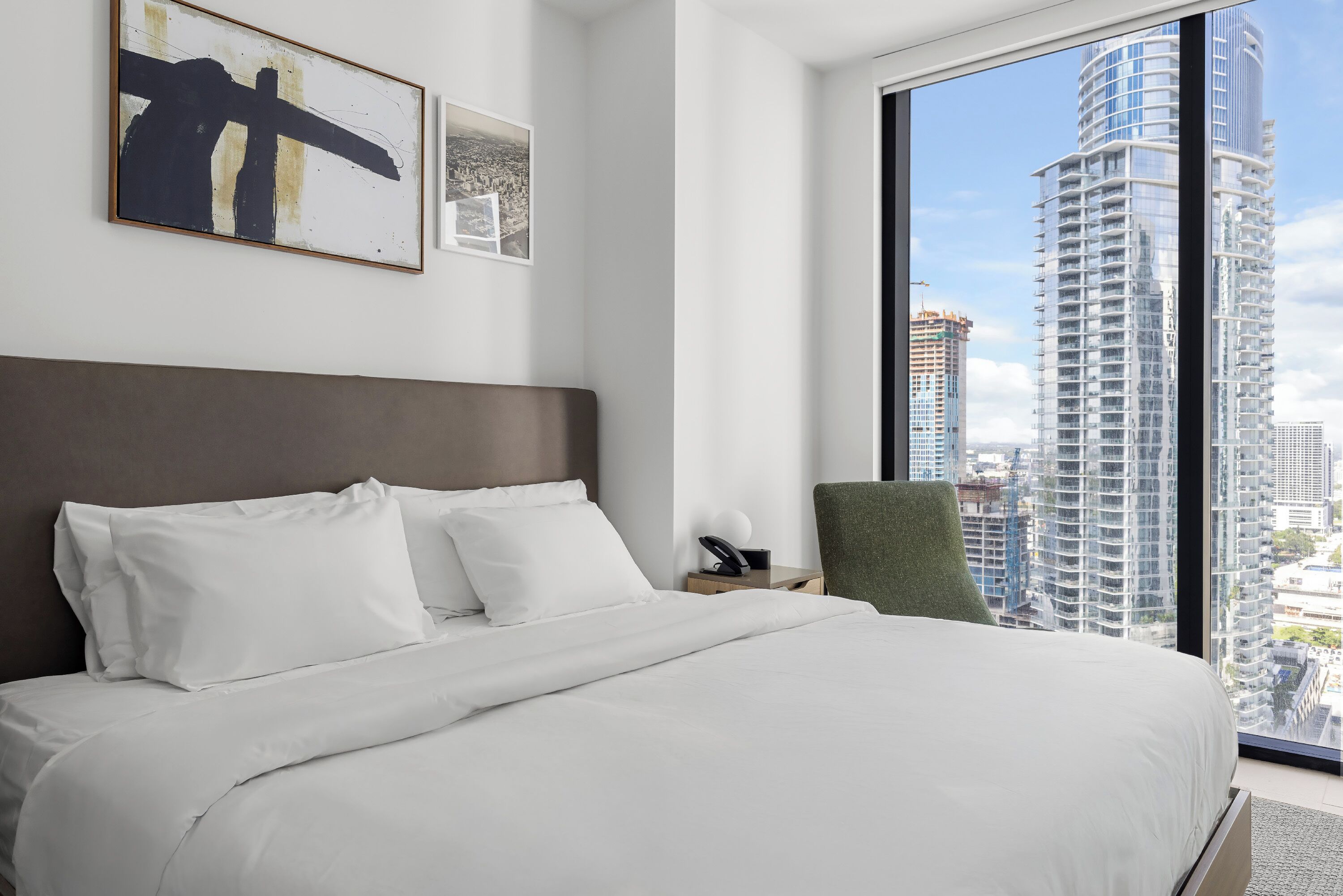 Een moderne hotelkamer met een groot bed, een groene stoel, een nachtkastje en een stadszicht door grote ramen.