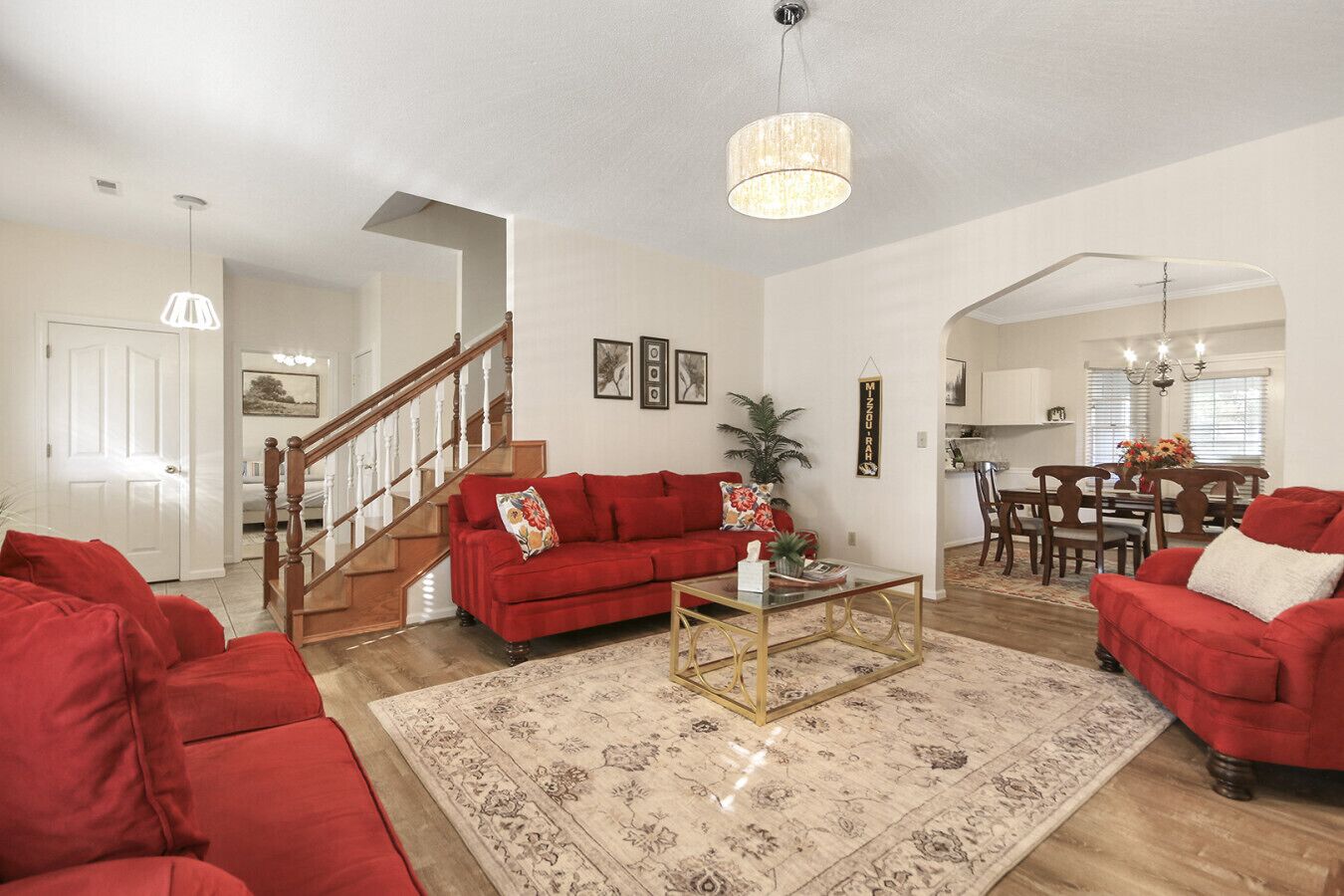 A spacious living room with a red sofa, a coffee table, and a chandelier. There is a staircase leading to another room, and a dining area with wooden furniture.