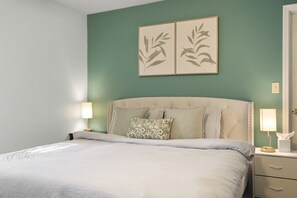 A neatly made bed with a beige headboard, two bedside lamps, and a green wall with two framed leaf designs.