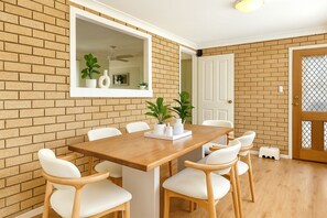 A dining area with a brick wall, a wooden table, white chairs, and a mirror above the table.