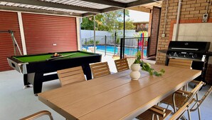 A pool table, a dining table with chairs, a grill, and a view of a swimming pool.