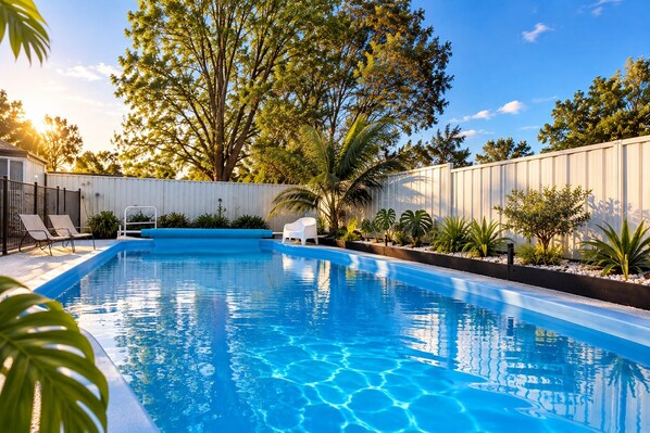A swimming pool with clear blue water, surrounded by a white fence, and shaded by trees.