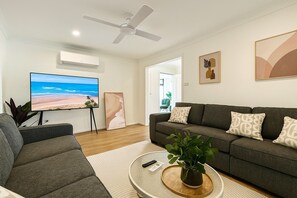 Un salon avec un canapé gris, un téléviseur à écran plat, un ventilateur de plafond et un tableau représentant une plage, encadré.