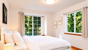 Um quarto com uma cama, uma mesa de cabeceira com abajur e janelas com cortinas.
