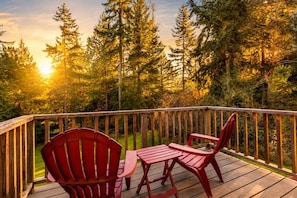 A wooden deck with a table and two chairs, overlooking a forest with tall trees and a sunset.